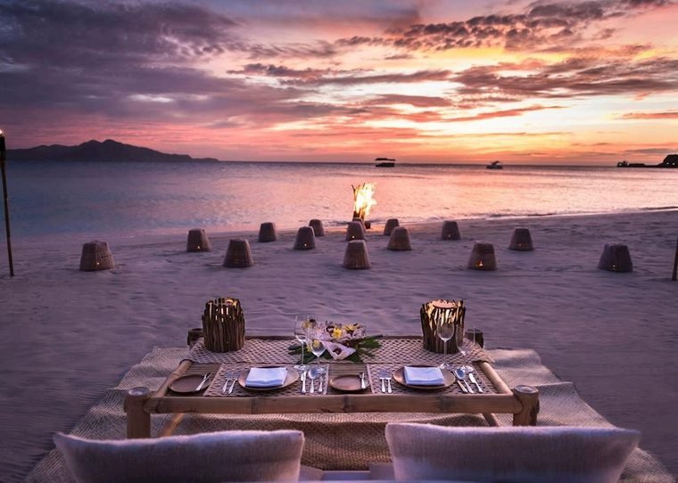 Low beach dining table set for dinner on the sand, ringed by candles under a pink sunset