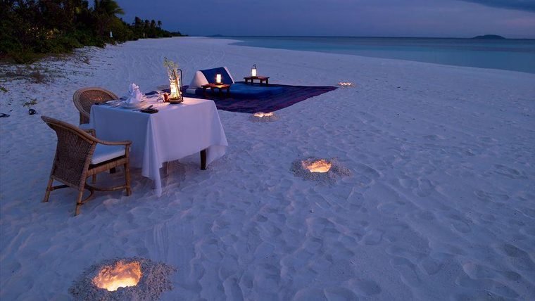 Private candlelit dinner table for two set on the white sand beside the water at dusk