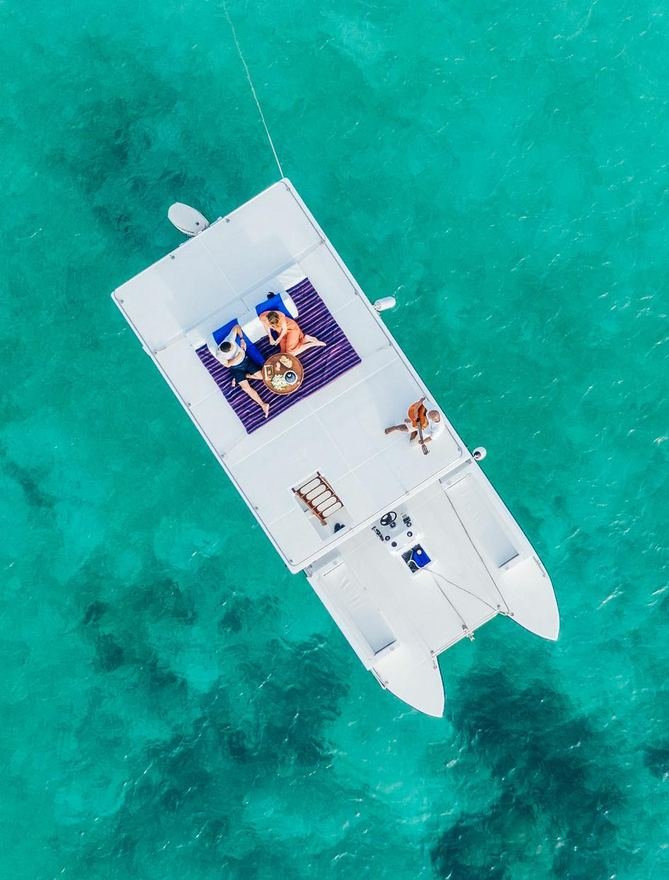 Aerial view of a couple relaxing on the deck of a catamaran anchored over turquoise reef