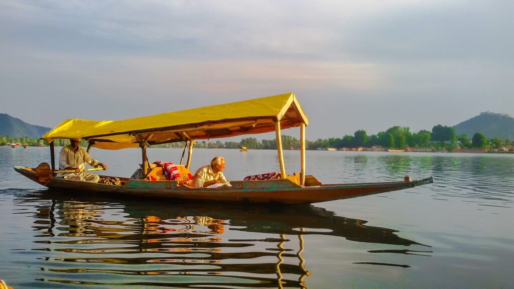 "A beautifully decorated Shikara boat floating on the serene waters of Dal Lake in Srinagar, Kashmir, with a backdrop of snow-capped Himalayas and charming houseboats. The tranquil ride offers stunning reflections, lush floating gardens, and a glimpse into the unique Kashmiri lifestyle."