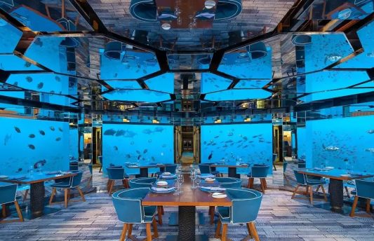 Luxury underwater restaurant at Anantara Kihavah Maldives with panoramic glass walls showcasing tropical fish, elegant dining setup with blue chairs, and mirrored ceiling reflecting marine life.